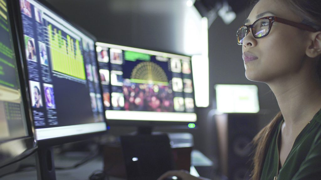 Close up stock image of a young asian woman sitting down at her desk where she’s surrounded by 3 large computer monitors displaying out of focus images of people as thumbnails, crowds, and graphs and scrolling text.