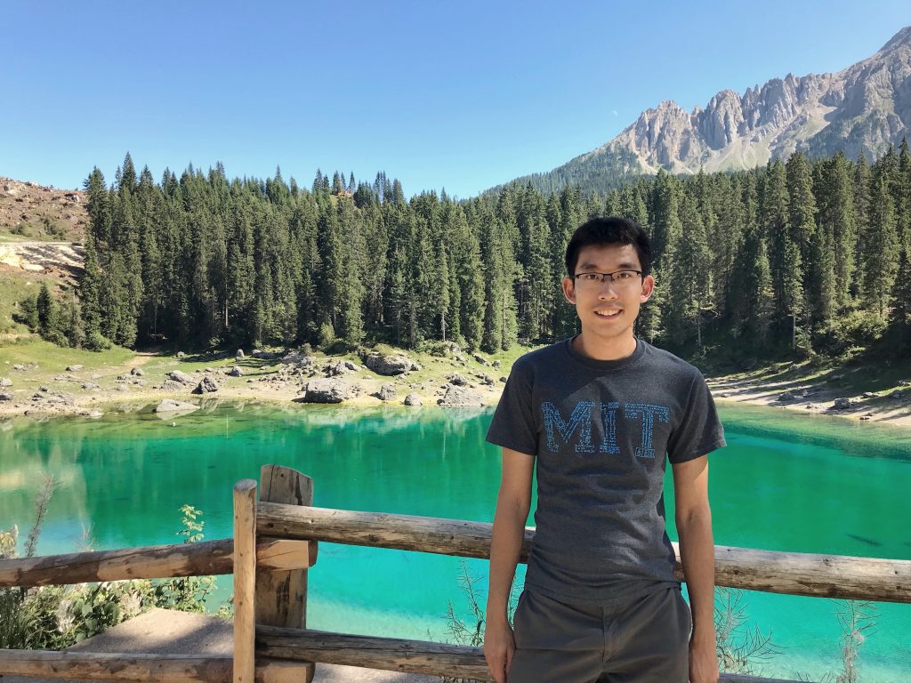 Minghao Qiu, wearing an MIT t-shirt, stands in front of a small green pond with trees and mountains in the background