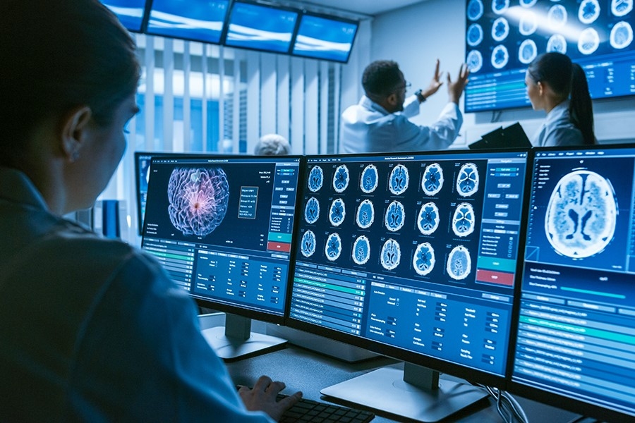 technician reviewing brain scans on a several monitors while colleagues look at scans on a light board